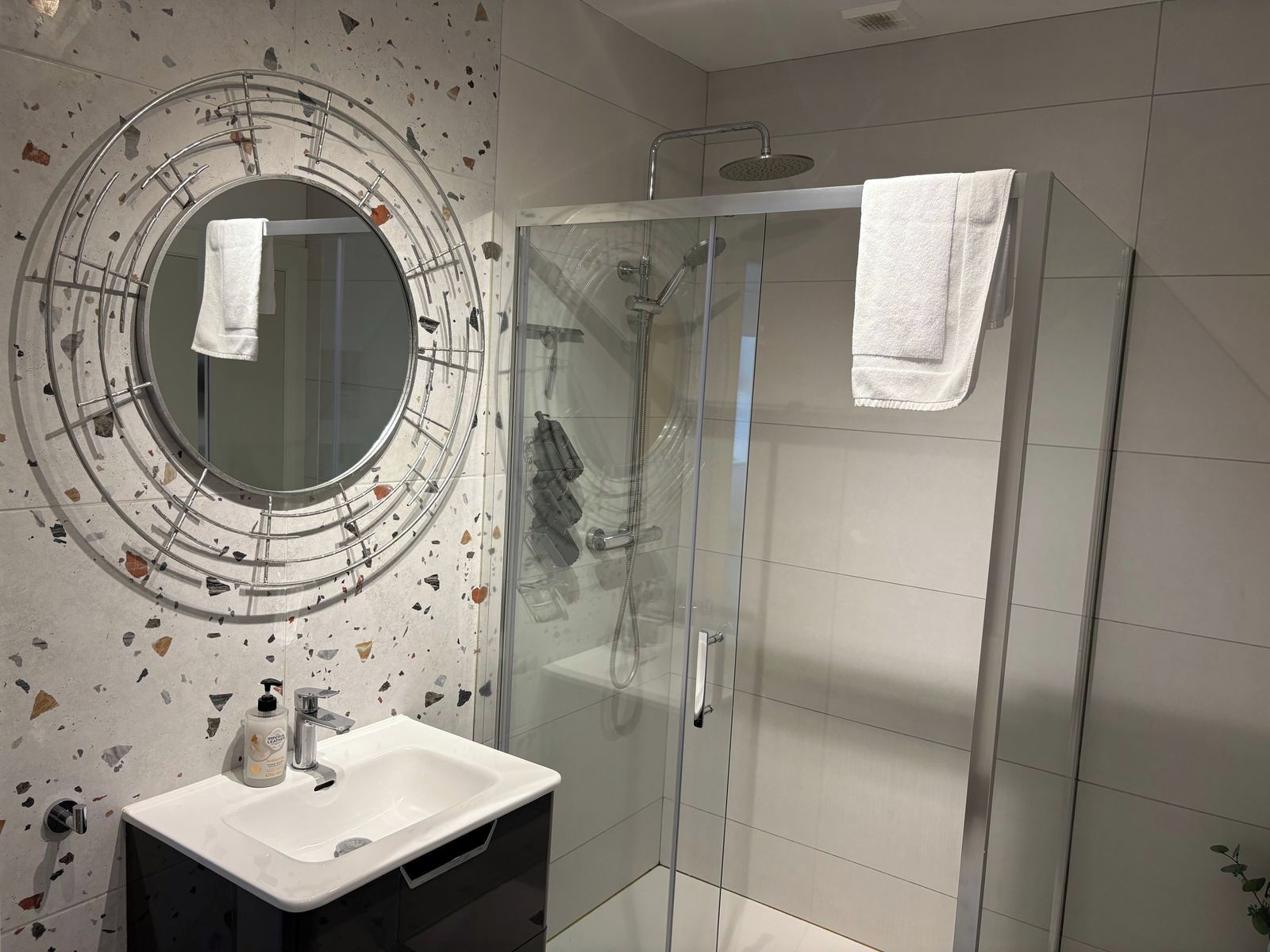 Une salle de bain moderne avec une douche à parois de verre, un grand miroir rond encadré, et un lavabo blanc agrémenté d’accessoires de toilette.