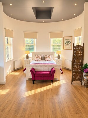 Quarto com cama, mesas de cabeceira, um banquinho, radiador e um painel decorativo.