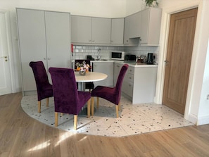 Una cocina moderna con armarios blancos, microondas y cafetera. Hay una mesa redonda con sillas moradas y un florero con flores.