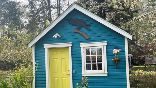 A small blue house with a yellow door, a window, and a bird sculpture on the roof.