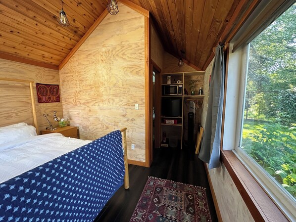 A bedroom with a bed, a nightstand, a desk, a chair, and a window with a view of greenery.