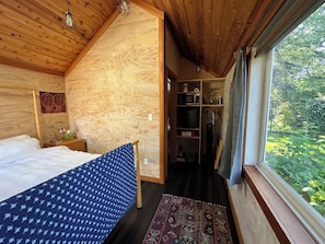 Un dormitorio con cama, mesita de noche, escritorio, silla y ventana con vistas al verdor.