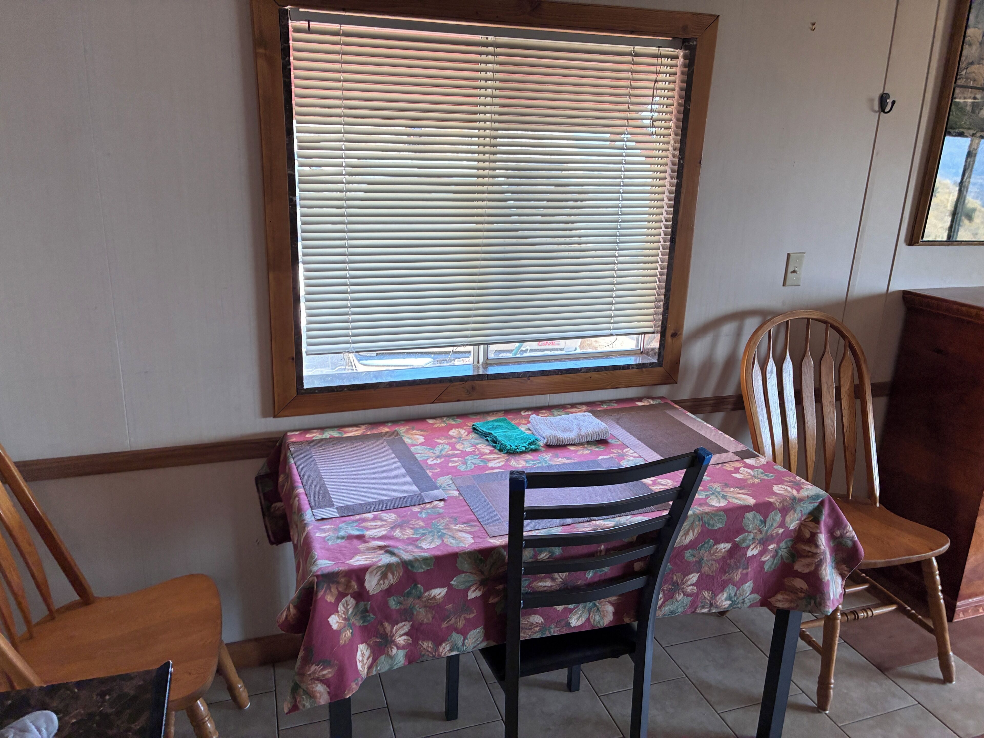 Un coin repas avec une table recouverte d’une nappe à motifs floraux, deux chaises en bois et une fenêtre avec des stores.