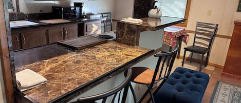 Una cocina con encimera de mármol, armarios de madera, cafetera y hervidor. Hay dos taburetes de bar y un otoman azul.