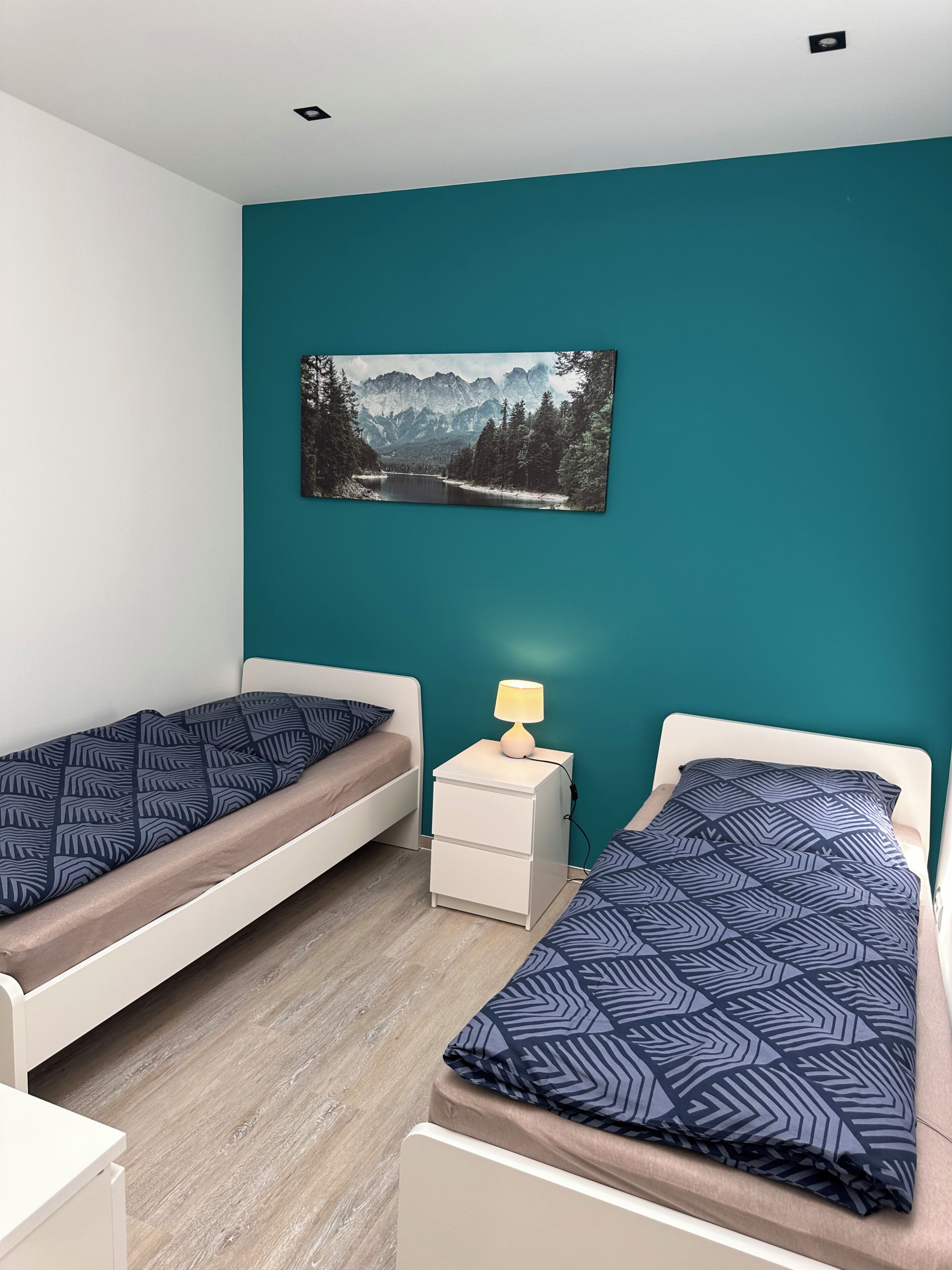 Ein Schlafzimmer mit zwei Einzelbetten, einem Nachttisch mit Lampe und einem Wandbild einer Berglandschaft.