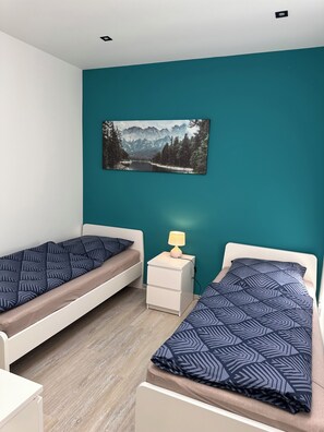 Ein Schlafzimmer mit zwei Einzelbetten, einem Nachttisch mit Lampe und einem Wandbild einer Berglandschaft.