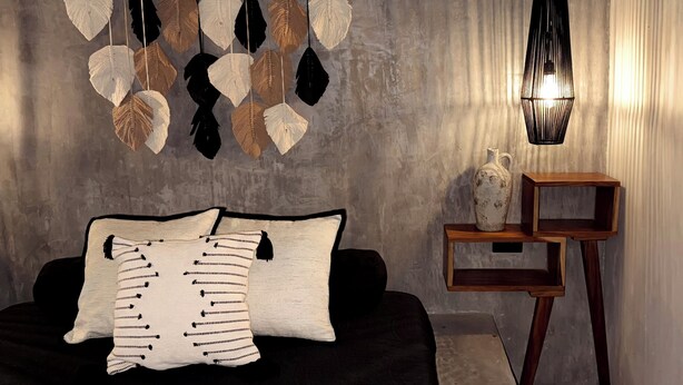 Una habitación con una cama, un adorno de hoja colgante, un jarrón sobre una mesita de madera y una lámpara fijada a la pared.