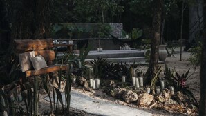 Un banco con cojines, rodeado de rocas y plantas, con velas y un sendero que lleva a una estructura al fondo.
