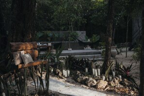 Un banc agrémenté de coussins, entouré de rochers et de plantes, avec des bougies et un sentier menant à une construction visible au loin.
