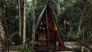 Una cabaña moderna de madera con forma triangular, rodeada de un bosque denso, con grandes ventanales y suelo de grava.