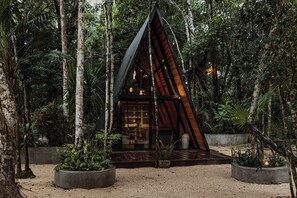 Une cabane moderne en bois, de forme triangulaire, entourée d’une forêt dense, dotée de grandes fenêtres et d’un sol recouvert de gravier.