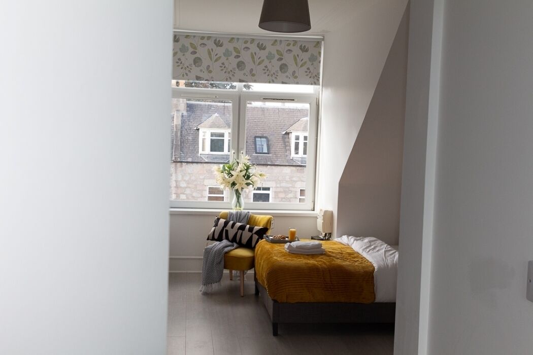Een slaapkamer met een bed, een stoel, een raam met uitzicht op gebouwen en een lamp.