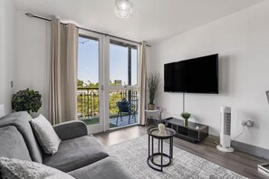 A modern living room with a grey sofa, a flat-screen TV, a standing fan, and a balcony with a view of greenery. - Central Apt - Balcony | Netflix | Walk to Bullring (Birmingham)