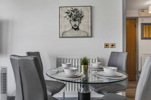 A modern dining area with a glass table, grey chairs, white plates, and a potted plant. - Central Apt - Balcony | Netflix | Walk to Bullring (Birmingham)