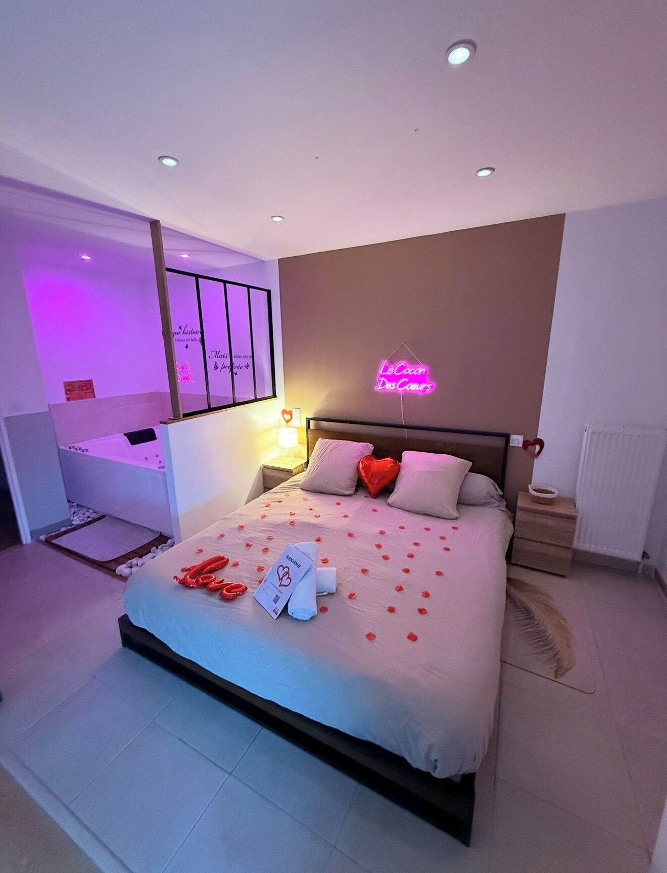 A hotel room with a bed, bedside tables, a nightstand, a wall-mounted lamp, and a neon sign.