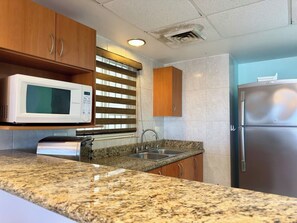 A kitchen with granite countertops, a stainless steel refrigerator, a microwave, and wooden cabinets. - Elegante Departamento en Costa Veleros (Mazatlán)