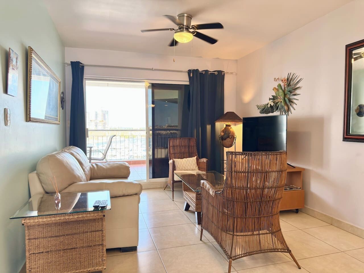 A living room with a sofa, armchairs, a glass coffee table, a ceiling fan, and a view through a glass door to an outdoor balcony.
