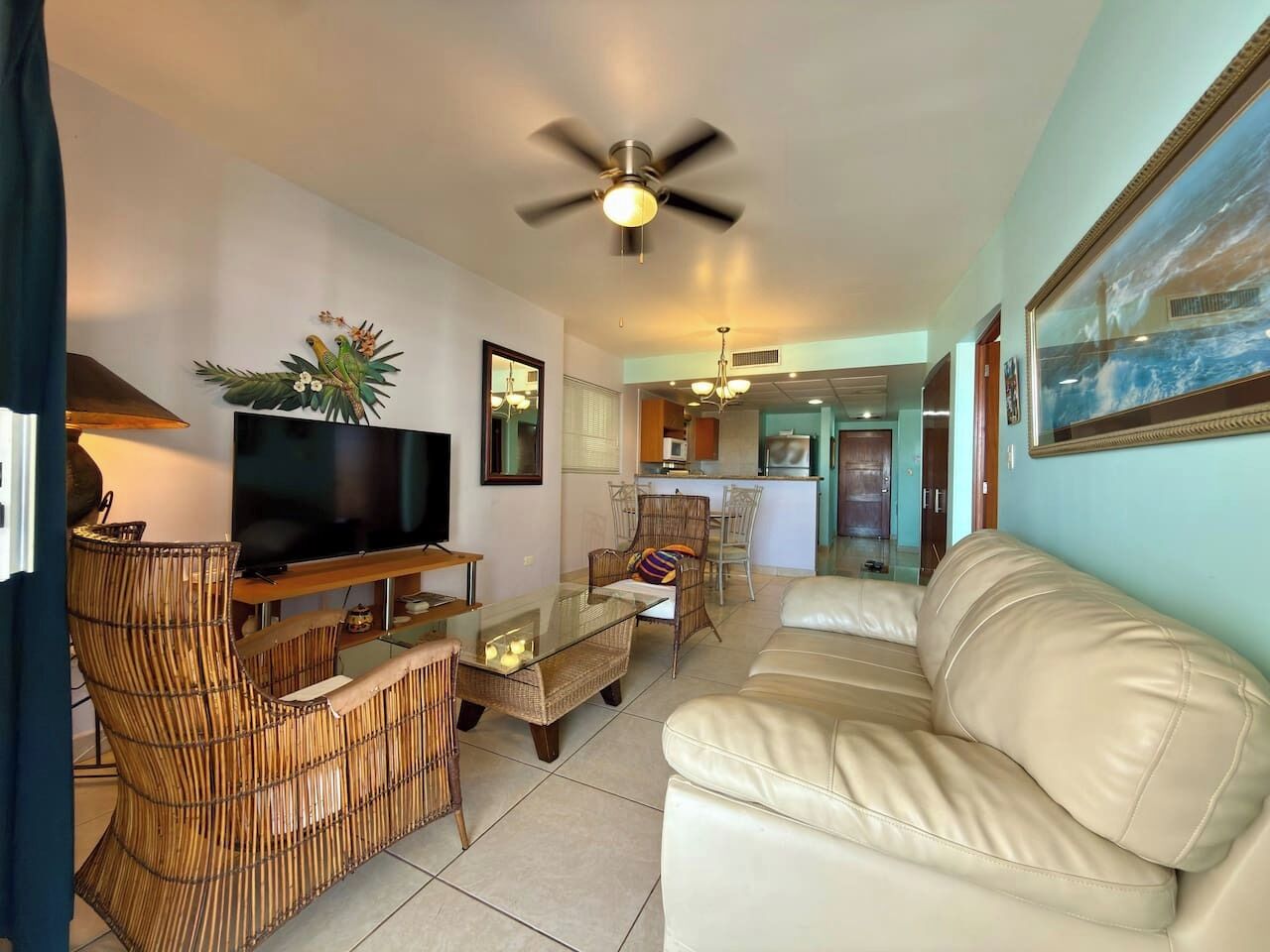 A living room with a television, wicker furniture, a ceiling fan, and a view into the kitchen.