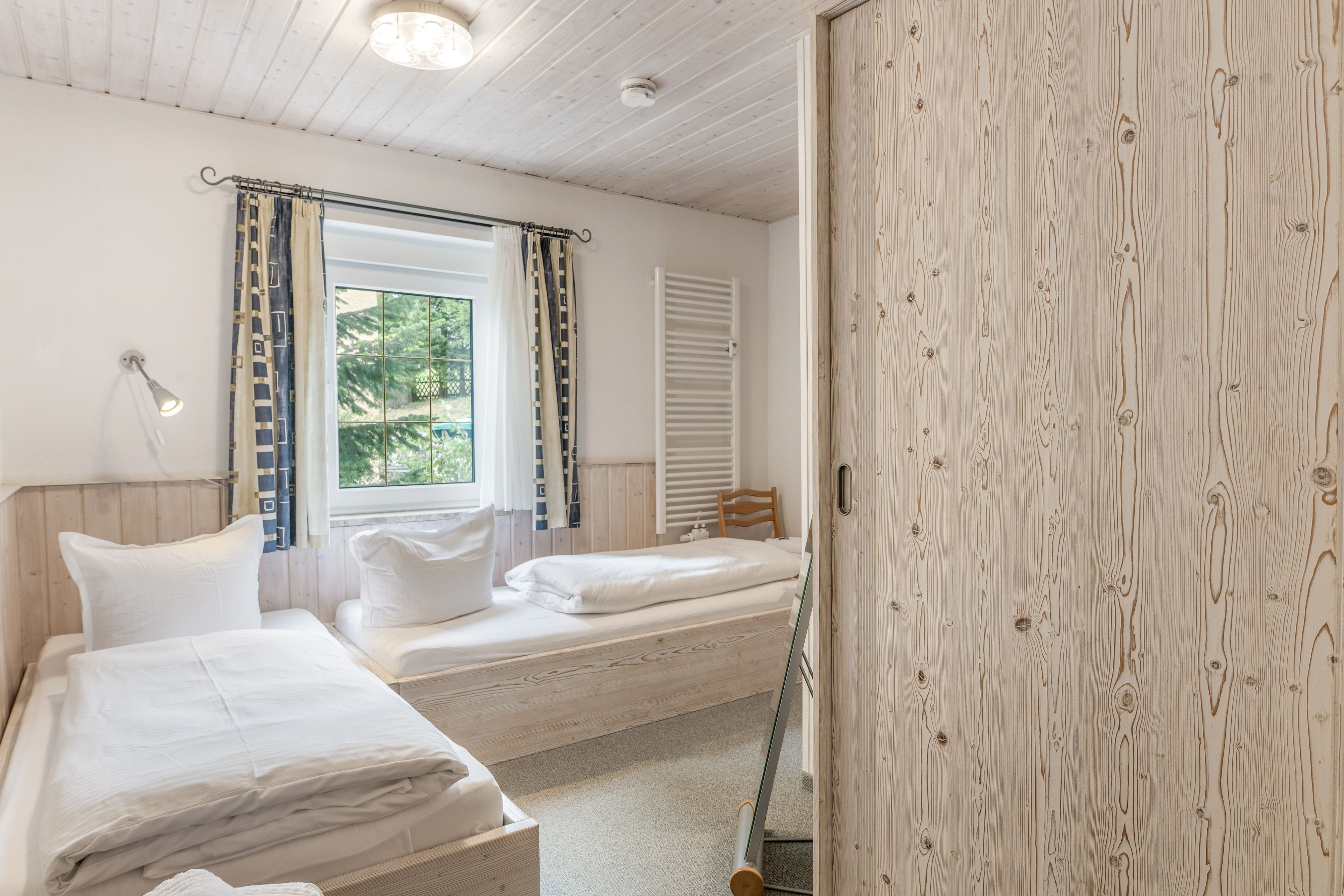 Quarto com duas camas, janela com cortinas, luminária de cabeceira e guarda-roupa de madeira.