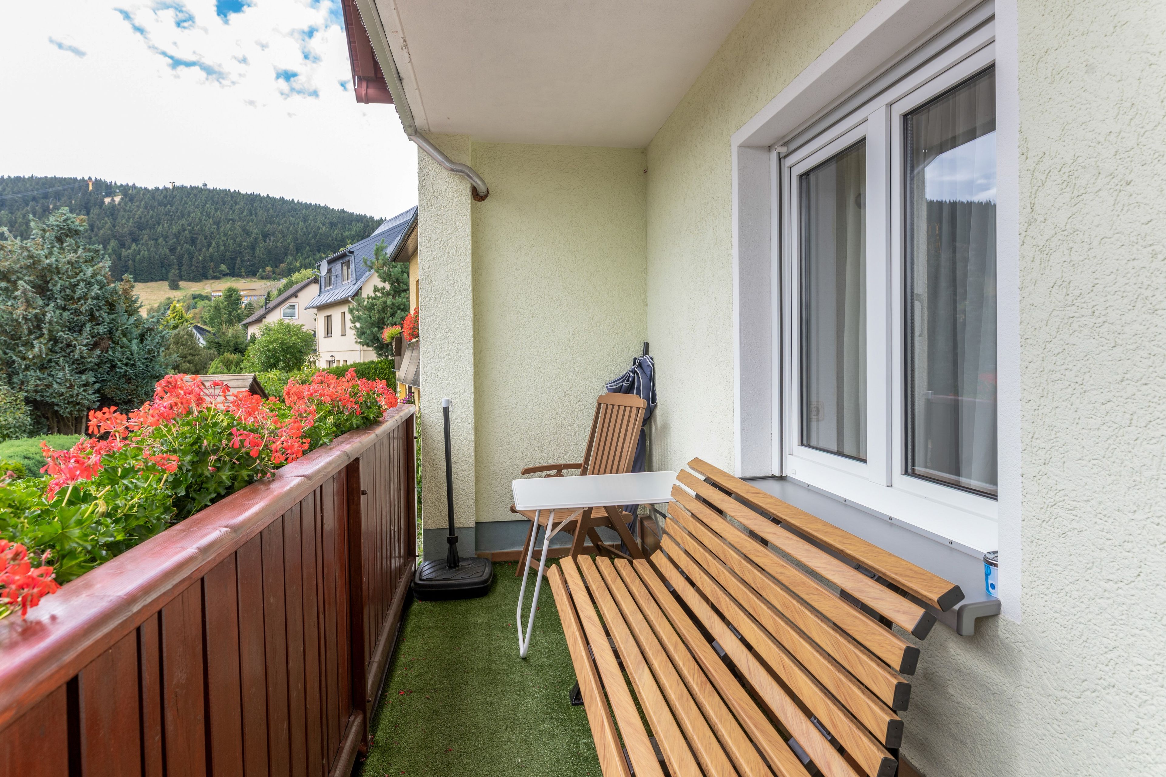 A balcony with a wooden bench, a table, and a chair. There is a window with curtains, and a view of a green landscape with trees and buildings in the distance.