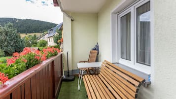 A balcony with a wooden bench, a table, and a chair. There is a window with curtains, and a view of a green landscape with trees and buildings in the distance.