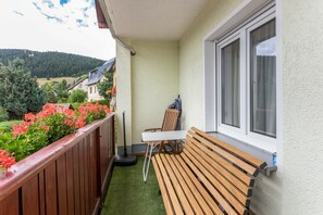 A balcony with a wooden bench, a table, and a chair. There is a window with curtains, and a view of a green landscape with trees and buildings in the distance. - Apartment "Ferienwohnungen Oberwiesenthal" with Mountain View, Balcony, and Wi-Fi (Oberwiesenthal)