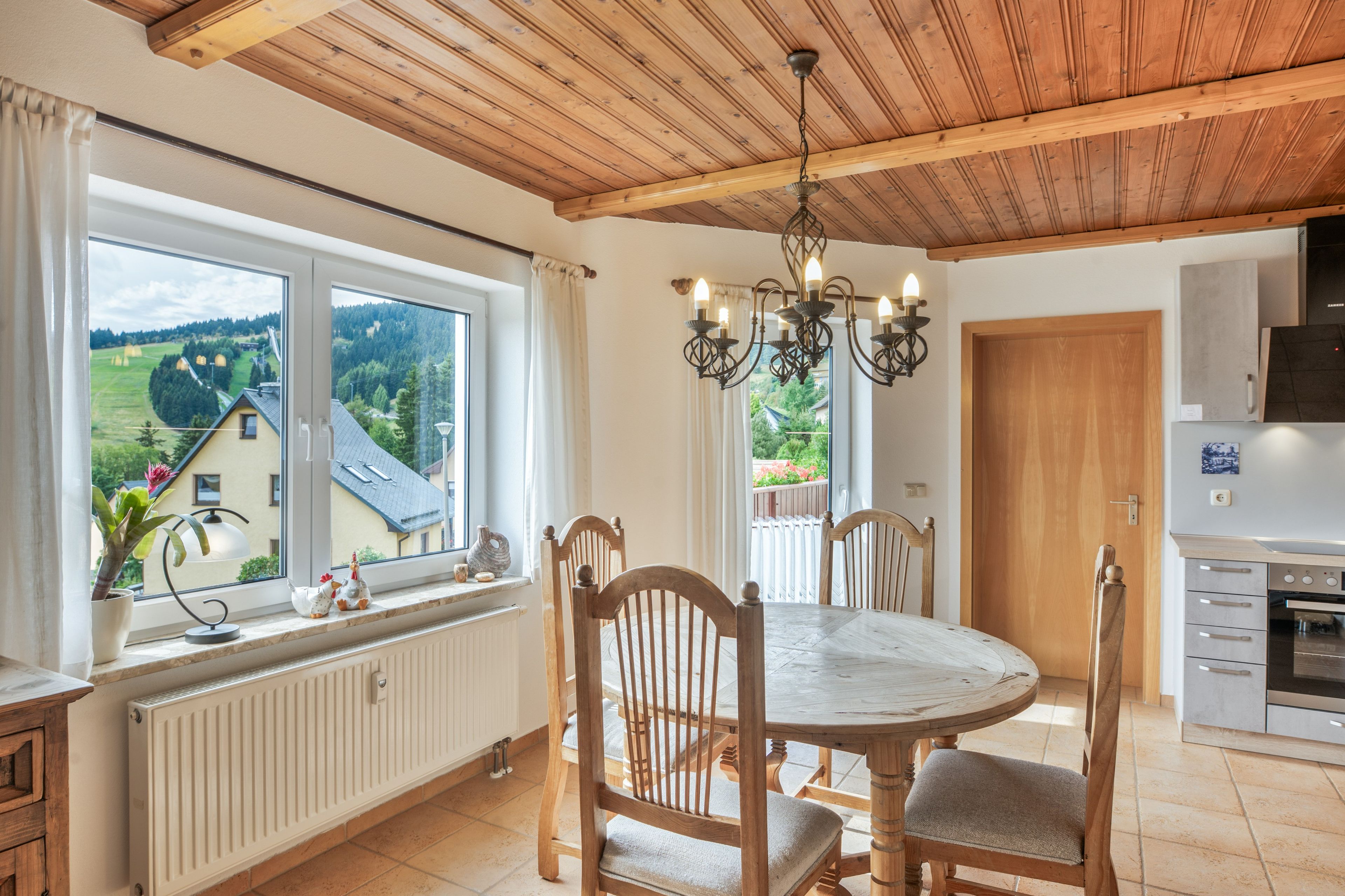 Espaço de refeições com mesa redonda e cadeiras, vista para o exterior através de amplas janelas, teto de madeira e área de cozinha com fogão e armários.