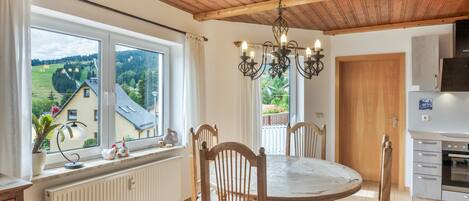 Een eetruimte met een ronde tafel en stoelen, uitzicht op het buitengebied door grote ramen, een houten plafond en een keuken met een fornuis en kastjes.