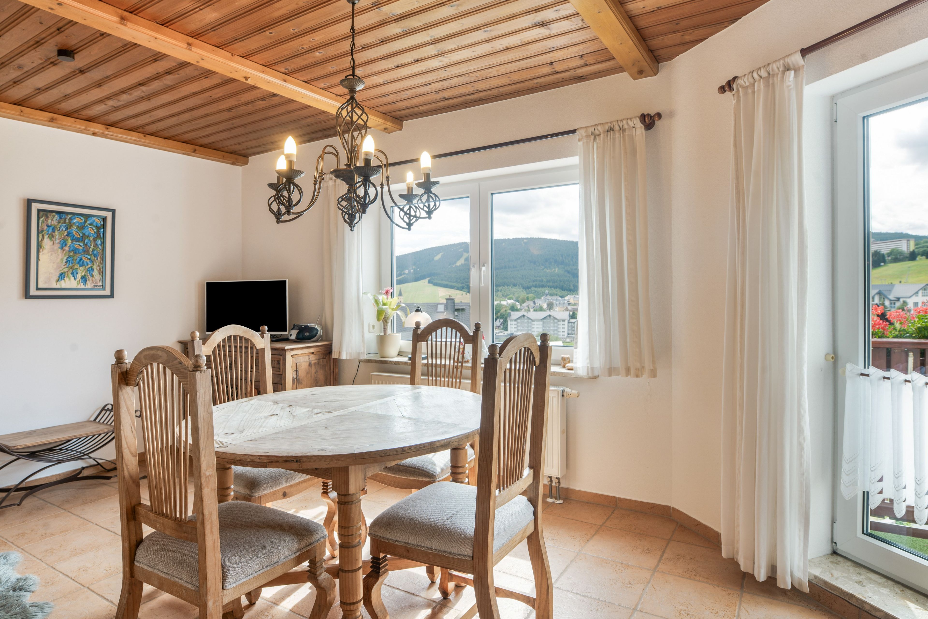A dining area with a round table and wooden chairs, a chandelier, a television, and a window with a view of a town and mountains.