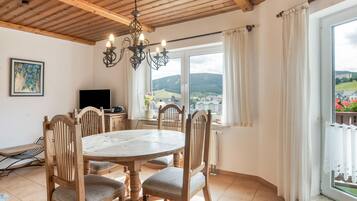 A dining area with a round table and wooden chairs, a chandelier, a television, and a window with a view of a town and mountains.