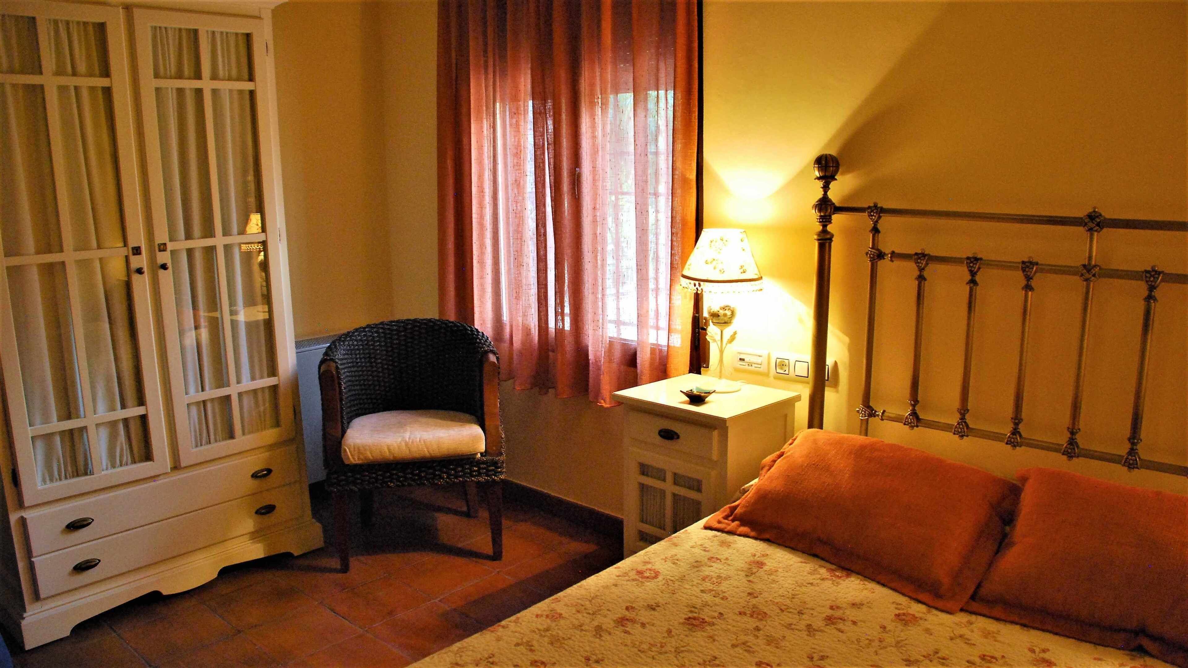 Une chambre à coucher comprenant un lit, une chaise, une table de chevet, une lampe, une armoire et une fenêtre avec des rideaux.