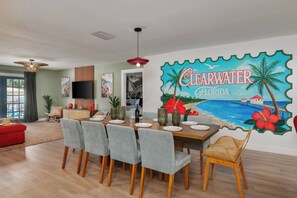 A dining area with a table set for four, surrounded by grey upholstered chairs. A large wall mural depicts a beach scene with the words 'Clearwater Florida.'
