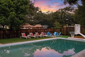 A backyard with a swimming pool, lounge chairs, a basketball hoop, and a wooden fence.