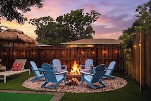 A backyard with a fire pit, four Adirondack chairs, a thatched umbrella, and a wooden fence.