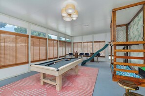 A pool table with billiard balls, a pool cue, and a green slide in a room with large windows and a wooden ladder.