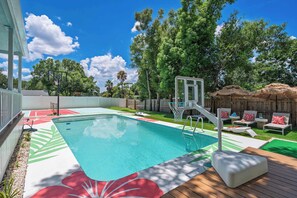 A swimming pool with a basketball hoop, surrounded by a wooden deck and lounge chairs.