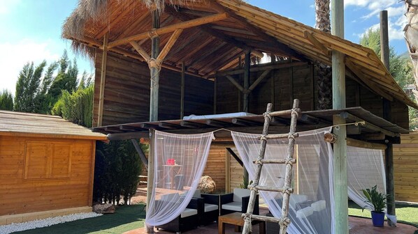 Une structure en bois avec un toit de chaume, une échelle et un espace de repos couvert, équipé d’une table et de chaises.