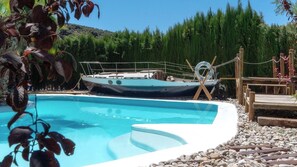 Une piscine avec un bateau amarré au bord, entourée d’une terrasse en bois et d’une rive rocheuse.