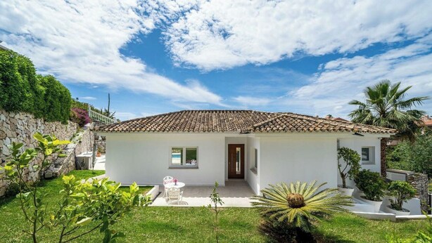 Uma casa térrea com telhado de telhas, parede de pedra e um pequeno jardim com uma mesa e cadeiras brancas.
