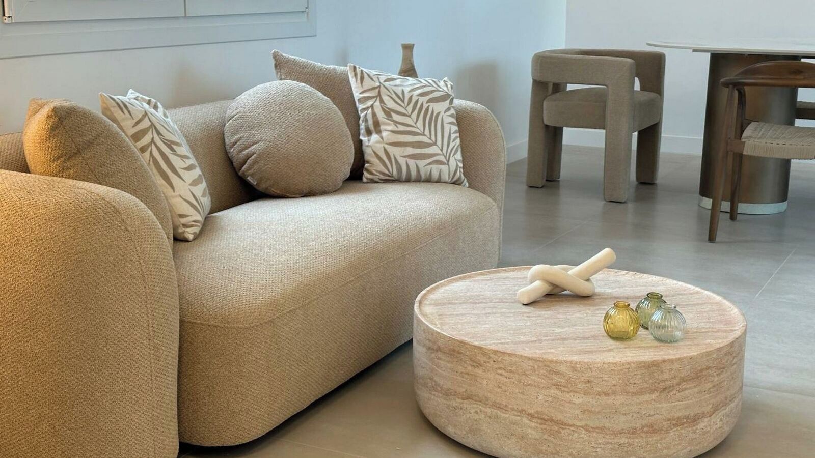 A modern living room with a beige sofa, patterned cushions, a round wooden coffee table with decorative items, and a dining area in the background.