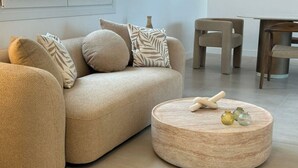 Uma sala de estar moderna com um sofá bege, almofadas estampadas, uma mesa de centro redonda de madeira com objetos decorativos, e uma área de jantar ao fundo.