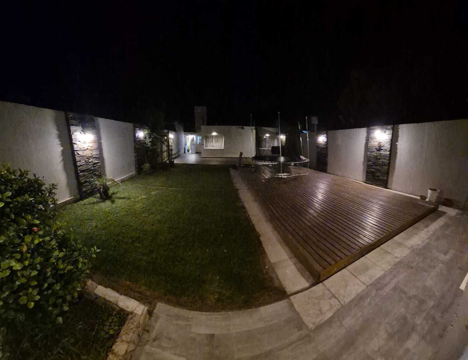 Une vue nocturne d’une cour avec une terrasse en bois, une petite pelouse et un chemin en pierre.