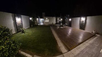 Une vue nocturne d’une cour avec une terrasse en bois, une petite pelouse et un chemin en pierre.