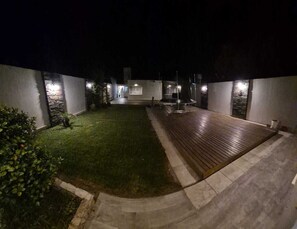 A nighttime view of a courtyard with a wooden deck, a small lawn, and a stone pathway.