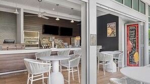 Ein moderner Barbereich mit weißen Korbstühlen und Tischen, einer Bar mit verschiedenen Flaschen und einer Kreidetafel mit der Speisekarte an der Wand.
