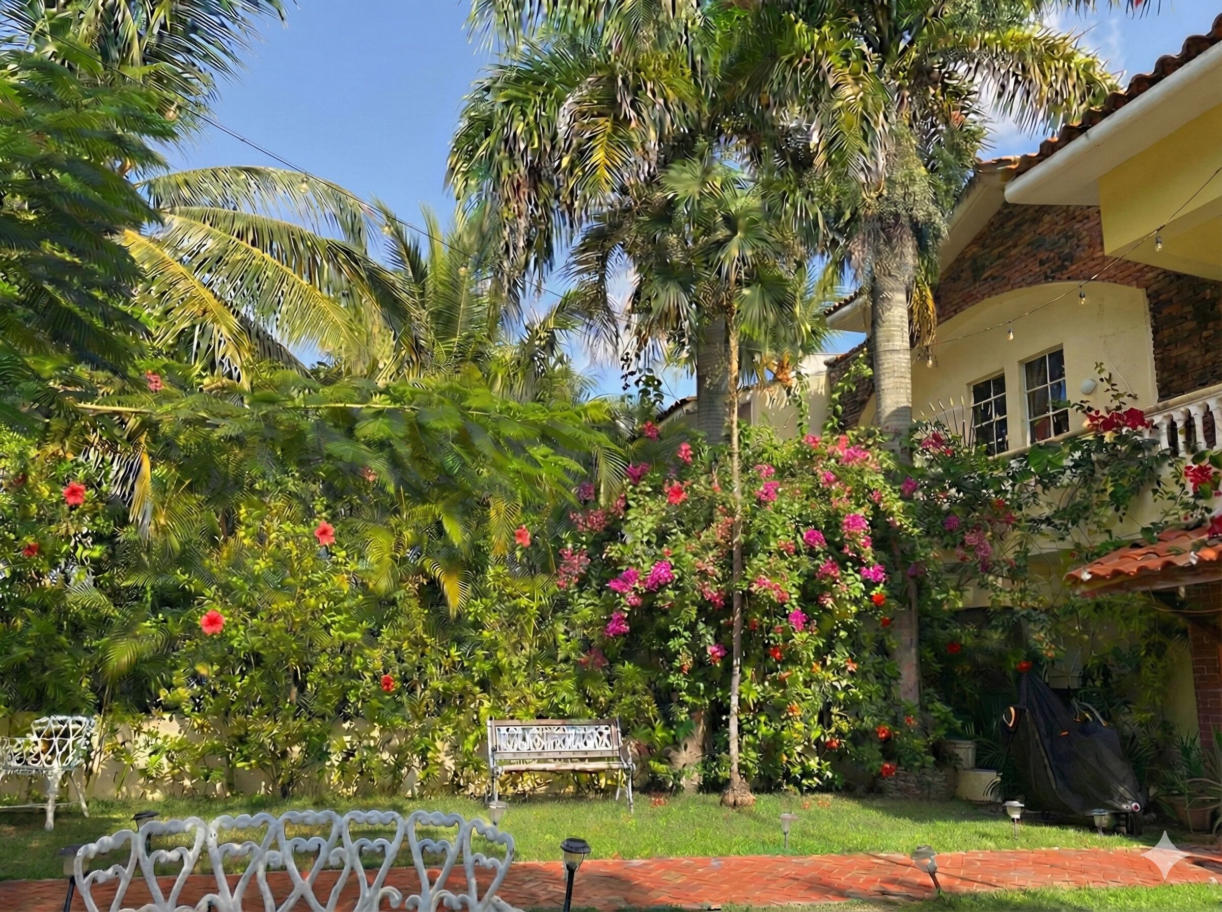 A garden with palm trees, flowering plants, a bench, and a building with a tiled roof.