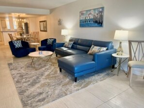A living room with a blue sofa, a coffee table, and a rug. There is a dining area in the background. - Beautiful Oceanfront 2/2 Condo, Private Balcony, Beachfront pool, No-Drive Beach (Daytona Beach)