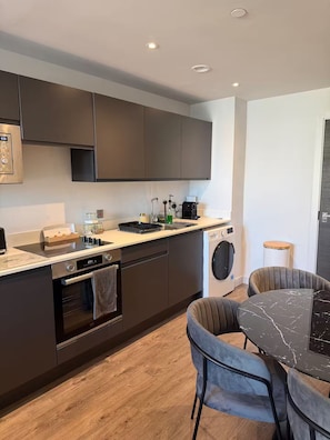 A modern kitchen with dark cabinets, a built-in oven, a dishwasher, and a coffee machine. - Apartment 1 Bedroom (Salford)