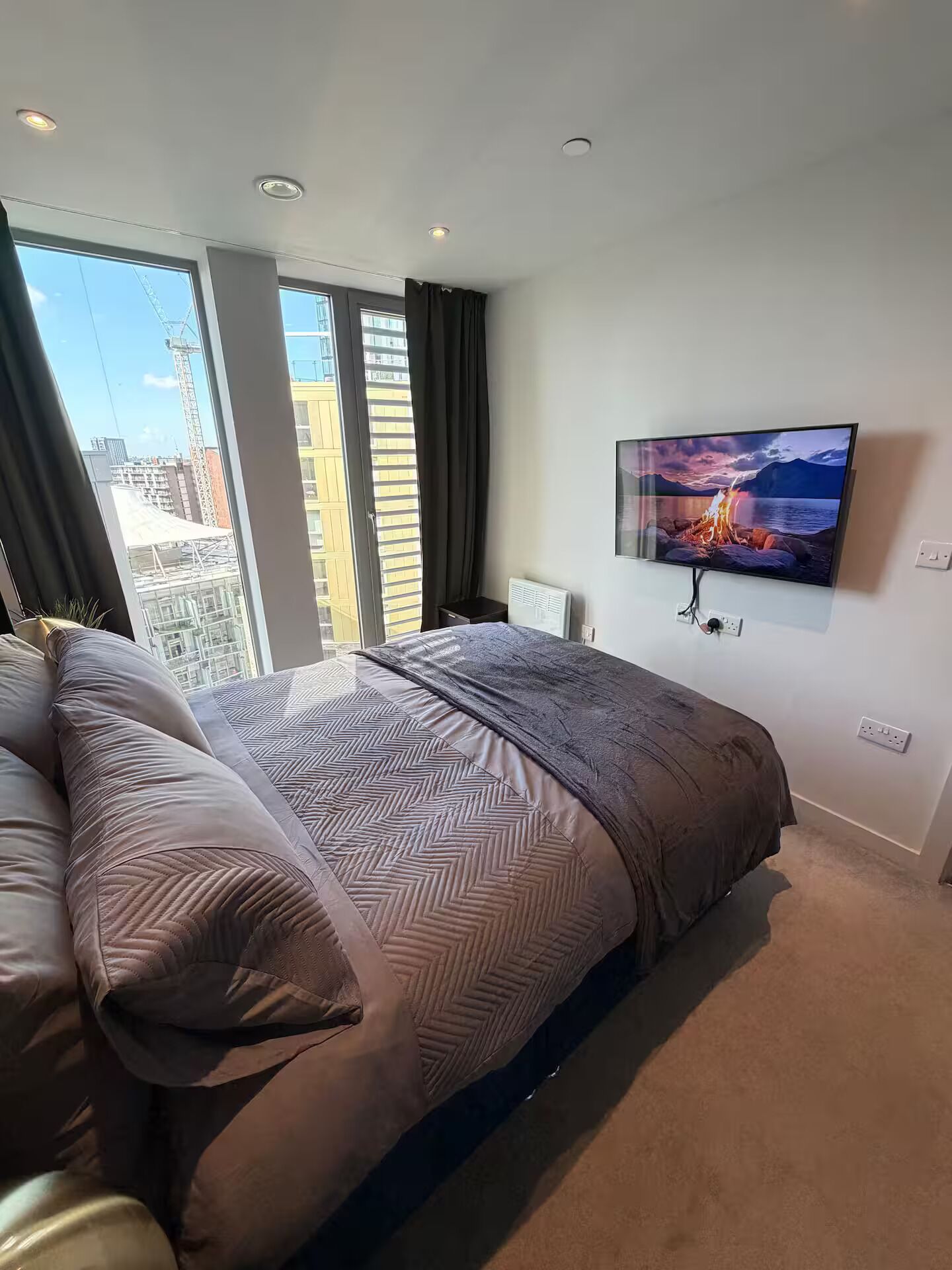 Un dormitorio moderno con una cama grande, un televisor montado en la pared y vistas de la ciudad a través de la ventana.