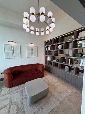 Ein modernes Wohnzimmer mit einem roten Sofa, einem grauen Hocker, einer Deckenleuchte und einem Bücherregal voller verschiedener Gegenstände.